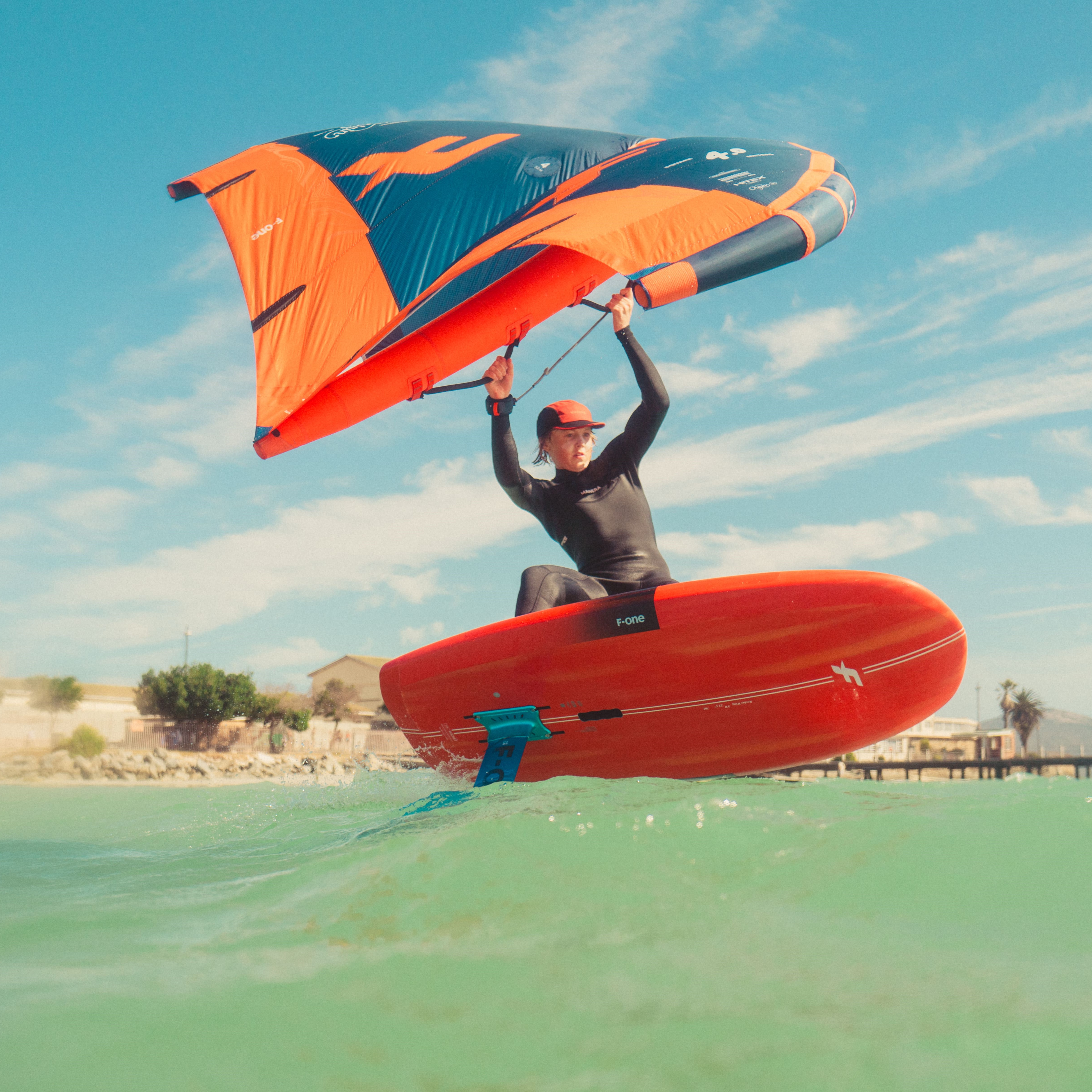Rideuse pratiquant le wingfoil avec l'aile F-One Origin V1 sur une mer calme, avec une plage en arrière-plan.