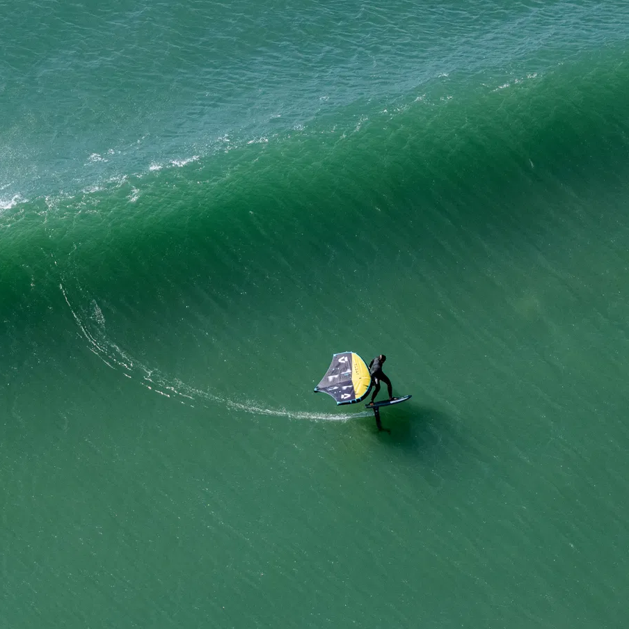 Rider en foil glissant sur une vague propre avec l’aile DUOTONE Float jaune et noire, configuration freefly, plan d’eau vert émeraude, angle aérien spectaculaire