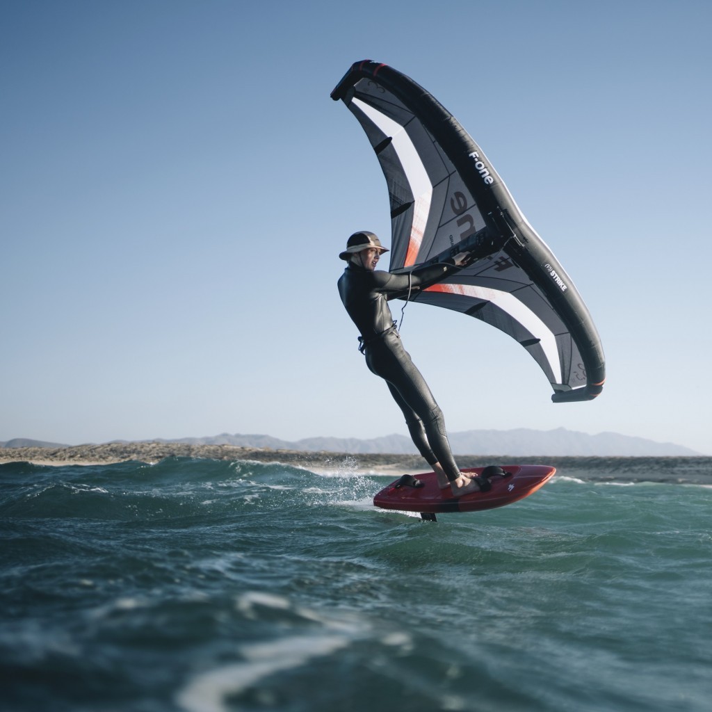 Rider en combinaison noire pratiquant le wingfoil avec une aile F-One Strike V5 sur une mer calme, avec un littoral en arrière-plan.