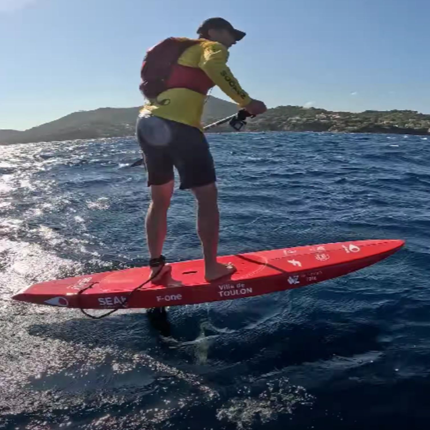 Rider en SUP foil F-One sur mer agitée, pagaie à la main, avec la côte méditerranéenne en arrière-plan